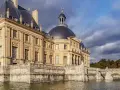 Le château de Vaux-le-Vicomte, majestueux édifice de pierre dorée, se reflète dans les eaux calmes, sous un ciel orageux.