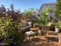 Aménagement terrasse jardin : salon de jardin en rotin, banquette confortable, table basse en bois, plantes luxuriantes et lanternes suspendues sous un ciel bleu.