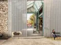 Un âne se tient devant la ferme de la Villette, une structure moderne contrastant avec un mur de pierre ancien. Deux personnes profitent du soleil sur des bancs.