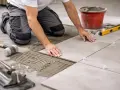 Un carreleur pose du carrelage au sol, appliquant la colle avec une truelle. Outils et seau de ciment à proximité.