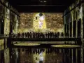 Un bassin des lumières projette "Le Baiser" de Klimt sur les murs d'une salle immersive. Des visiteurs contemplent l'œuvre depuis une passerelle.