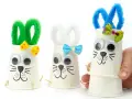 Trois adorables lapins de Pâques bricolage faits avec des gobelets en papier, des pompons et des yeux mobiles. Parfait pour une activité de Pâques !