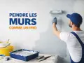 Un peintre en combinaison bleue utilise un rouleau pour repeindre un mur en blanc. Des pots de peinture et des pinceaux sont disposés sur une table.