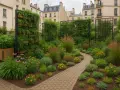 Un petit jardin de ville luxuriant avec un chemin sinueux, des plantes vertes, des graminées et des fleurs roses et jaunes, entouré d'immeubles.