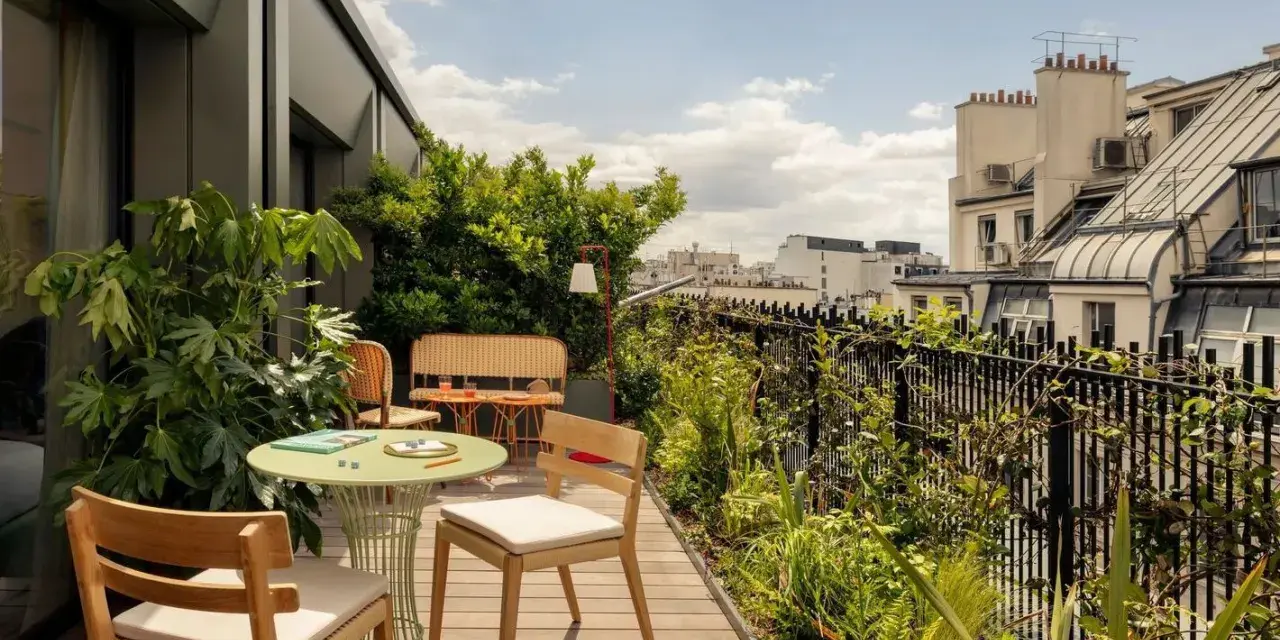 Aménagement balcon 10m2 : table ronde verte, chaises en bois, banquette en rotin, plantes luxuriantes et vue sur les toits de Paris.