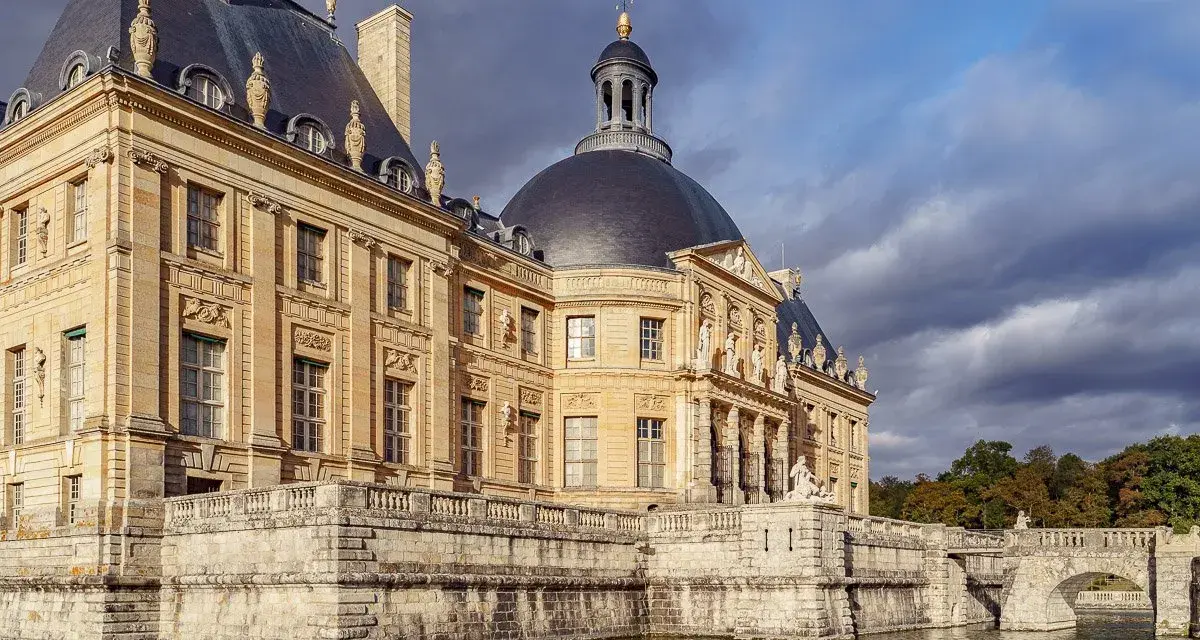 Le château de Vaux-le-Vicomte, majestueux édifice de pierre dorée, se reflète dans les eaux calmes, sous un ciel orageux.