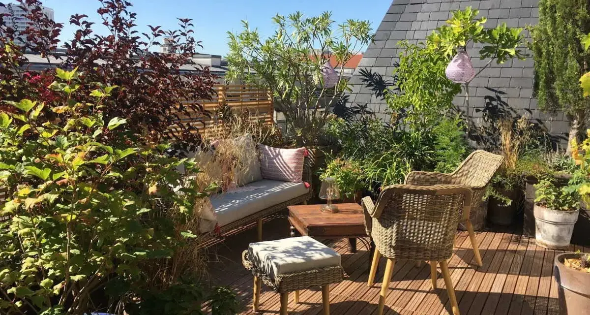 Aménagement terrasse jardin : salon de jardin en rotin, banquette confortable, table basse en bois, plantes luxuriantes et lanternes suspendues sous un ciel bleu.