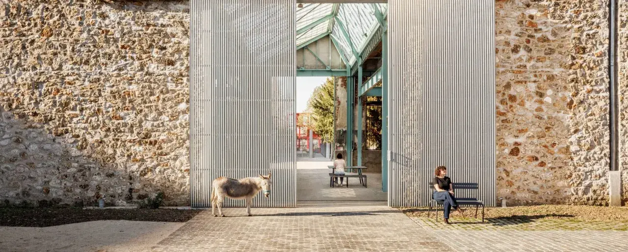 Un âne se tient devant la ferme de la Villette, une structure moderne contrastant avec un mur de pierre ancien. Deux personnes profitent du soleil sur des bancs.