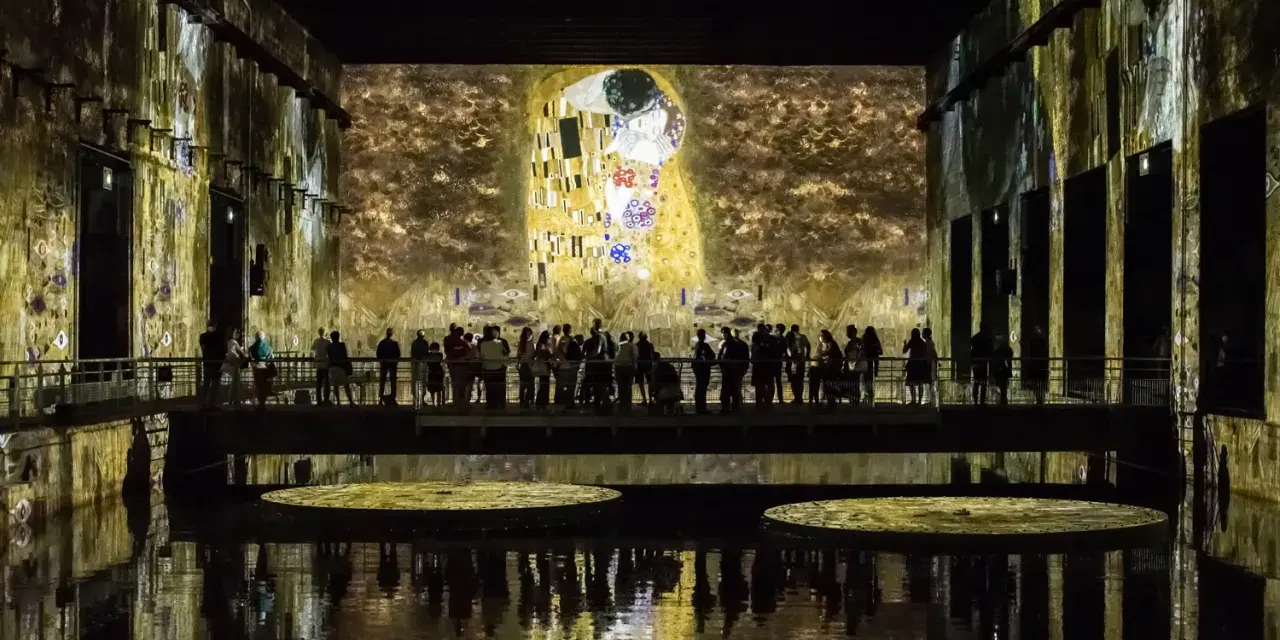 Un bassin des lumières projette "Le Baiser" de Klimt sur les murs d'une salle immersive. Des visiteurs contemplent l'œuvre depuis une passerelle.
