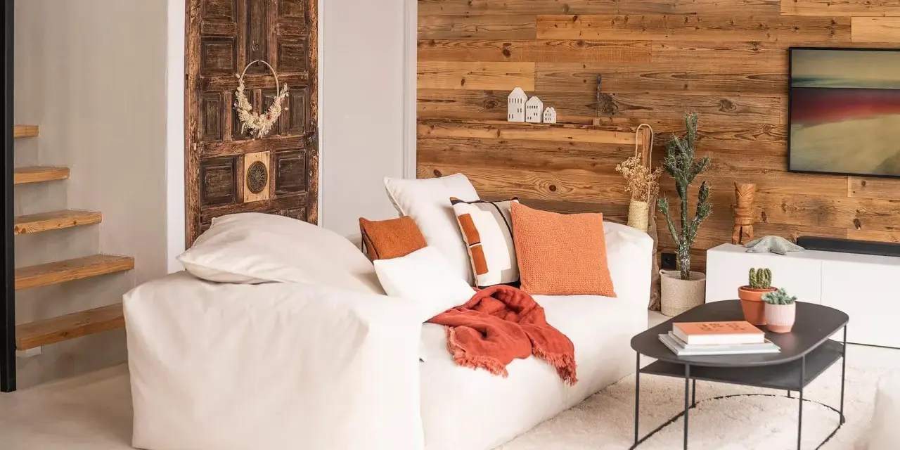 Un canapé blanc moelleux, des coussins orange et un plaid créent une deco petit salon cosy. Une porte ancienne et un mur en bois ajoutent du caractère.
