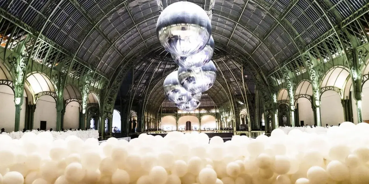 Installation de ballons blancs dans le Grand Palais, Paris. Des sphères argentées flottent sous la verrière, évoquant un musée des ballons.