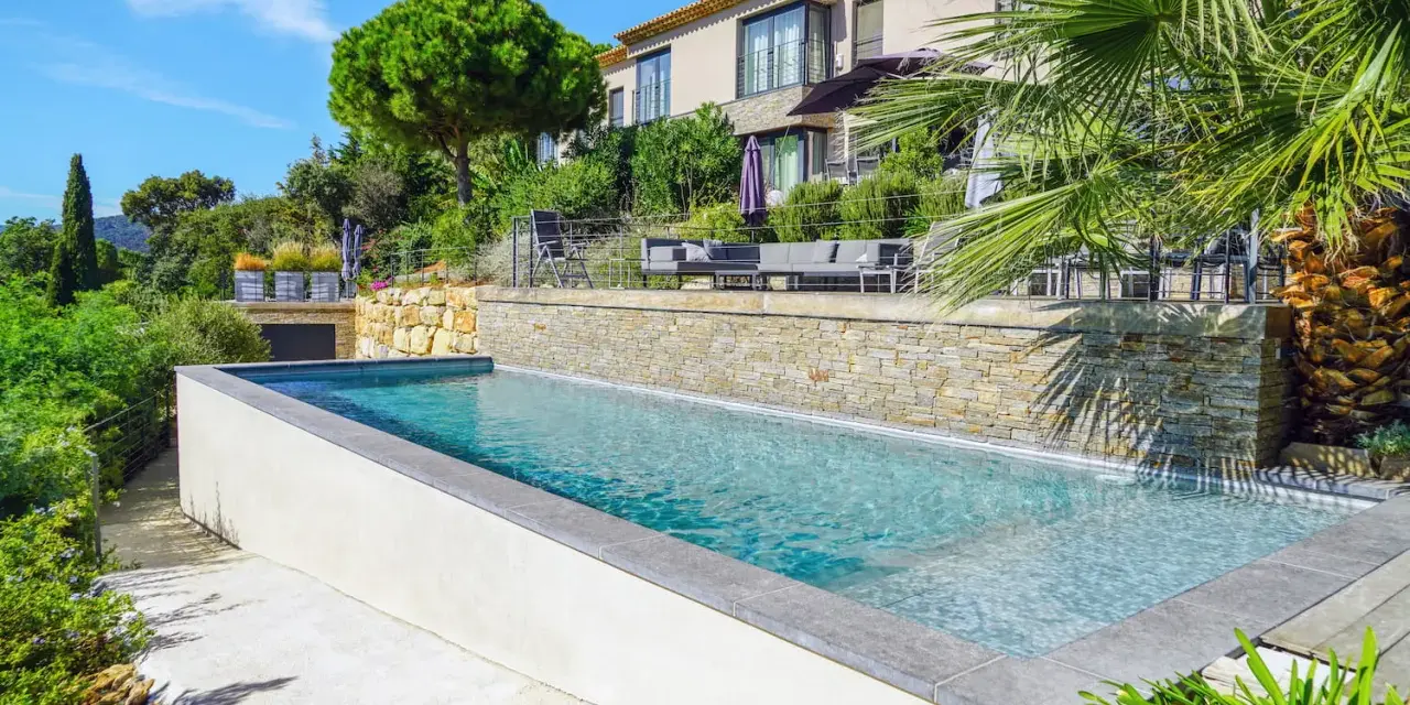 Magnifique piscine semi enterrée avec habillage en pierre, bordée par une terrasse et un jardin luxuriant, devant une villa moderne.
