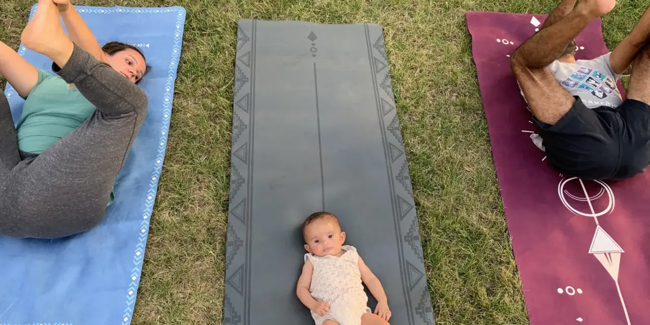 Un bébé sur un tapis de yoga, entouré de ses parents pratiquant le yoga. Une séance de yoga pour enfants en plein air.