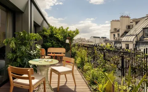 Aménagement balcon 10m2 : table ronde verte, chaises en bois, banquette en rotin, plantes luxuriantes et vue sur les toits de Paris.