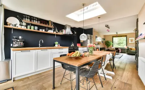 Cuisine moderne avec îlot central, parfait pour rénover vieille cuisine. Table en bois, chaises design, et mur noir avec étagères.
