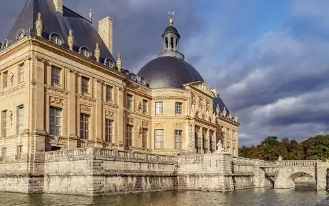 Le château de Vaux-le-Vicomte, majestueux édifice de pierre dorée, se reflète dans les eaux calmes, sous un ciel orageux.