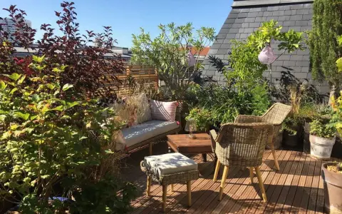Aménagement terrasse jardin : salon de jardin en rotin, banquette confortable, table basse en bois, plantes luxuriantes et lanternes suspendues sous un ciel bleu.