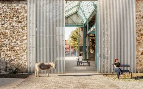 Un âne se tient devant la ferme de la Villette, une structure moderne contrastant avec un mur de pierre ancien. Deux personnes profitent du soleil sur des bancs.
