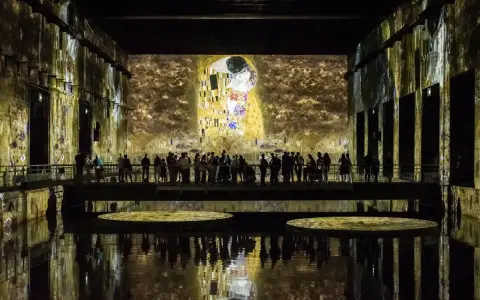 Un bassin des lumières projette "Le Baiser" de Klimt sur les murs d'une salle immersive. Des visiteurs contemplent l'œuvre depuis une passerelle.