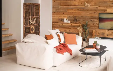 Un canapé blanc moelleux, des coussins orange et un plaid créent une deco petit salon cosy. Une porte ancienne et un mur en bois ajoutent du caractère.