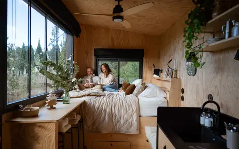 Un couple lit dans un lit douillet, profitant de l'aménagement ingénieux d'une tiny house avec vue sur la nature.