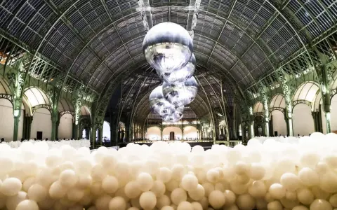 Installation de ballons blancs dans le Grand Palais, Paris. Des sphères argentées flottent sous la verrière, évoquant un musée des ballons.