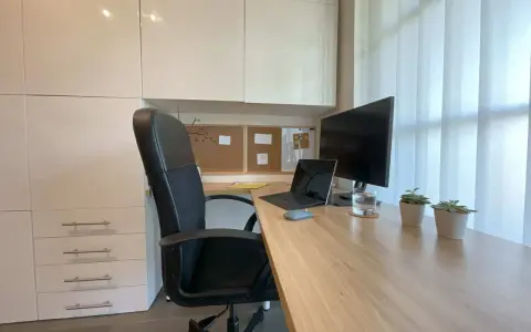 Bureau moderne avec fauteuil noir, bureau en bois clair, ordinateur portable, écran, plantes et stores blancs. Une belle idee deco bureau pour un espace fonctionnel et apaisant.