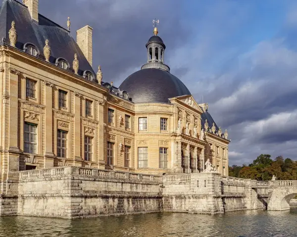 Le château de Vaux-le-Vicomte, majestueux édifice de pierre dorée, se reflète dans les eaux calmes, sous un ciel orageux.