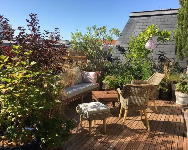 Aménagement terrasse jardin : salon de jardin en rotin, banquette confortable, table basse en bois, plantes luxuriantes et lanternes suspendues sous un ciel bleu.