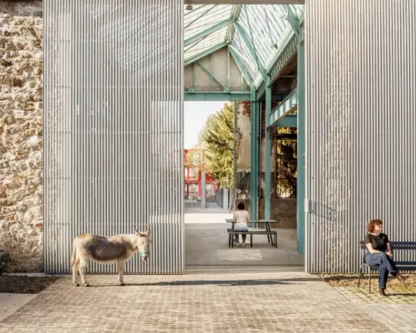 Un âne se tient devant la ferme de la Villette, une structure moderne contrastant avec un mur de pierre ancien. Deux personnes profitent du soleil sur des bancs.
