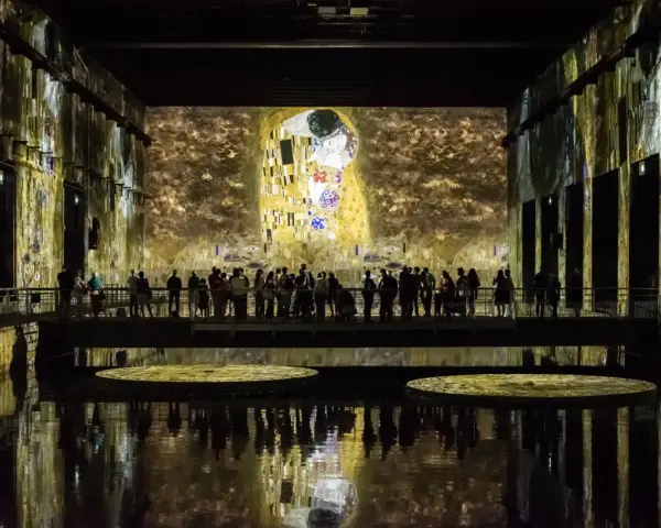 Un bassin des lumières projette "Le Baiser" de Klimt sur les murs d'une salle immersive. Des visiteurs contemplent l'œuvre depuis une passerelle.
