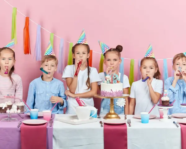 Six enfants joyeux célèbrent un anniversaire 11 ans, portant des chapeaux de fête et soufflant dans des cotillons devant un gâteau décoré.