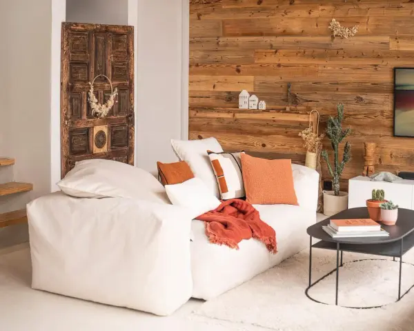 Un canapé blanc moelleux, des coussins orange et un plaid créent une deco petit salon cosy. Une porte ancienne et un mur en bois ajoutent du caractère.