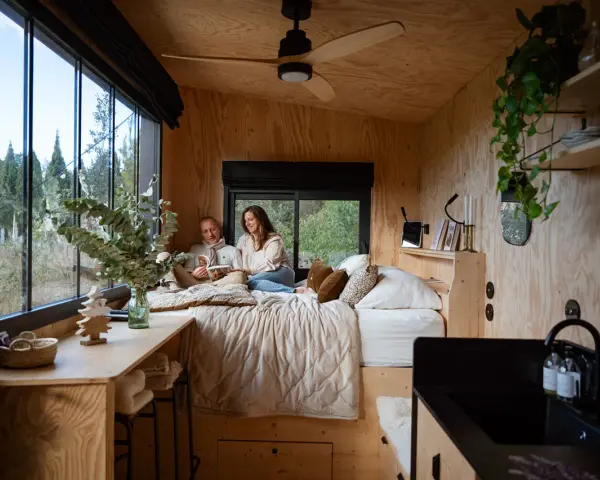 Un couple lit dans un lit douillet, profitant de l'aménagement ingénieux d'une tiny house avec vue sur la nature.