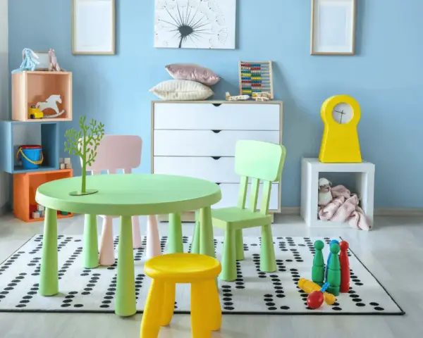 Salle de jeux lumineuse avec table verte, chaises colorées, jouets et peluche sur fond bleu clair.