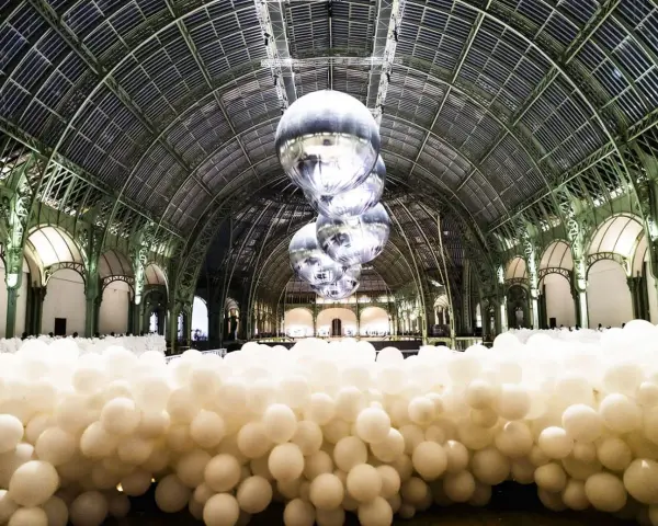 Installation de ballons blancs dans le Grand Palais, Paris. Des sphères argentées flottent sous la verrière, évoquant un musée des ballons.