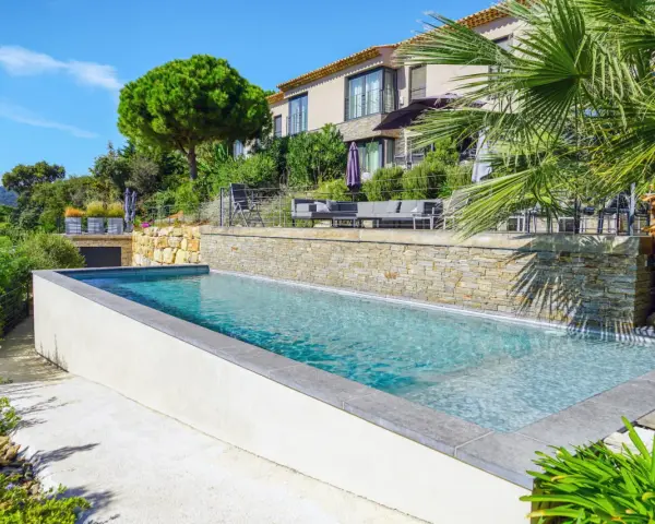 Magnifique piscine semi enterrée avec habillage en pierre, bordée par une terrasse et un jardin luxuriant, devant une villa moderne.
