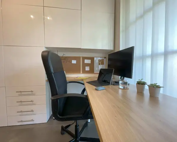 Bureau moderne avec fauteuil noir, bureau en bois clair, ordinateur portable, écran, plantes et stores blancs. Une belle idee deco bureau pour un espace fonctionnel et apaisant.