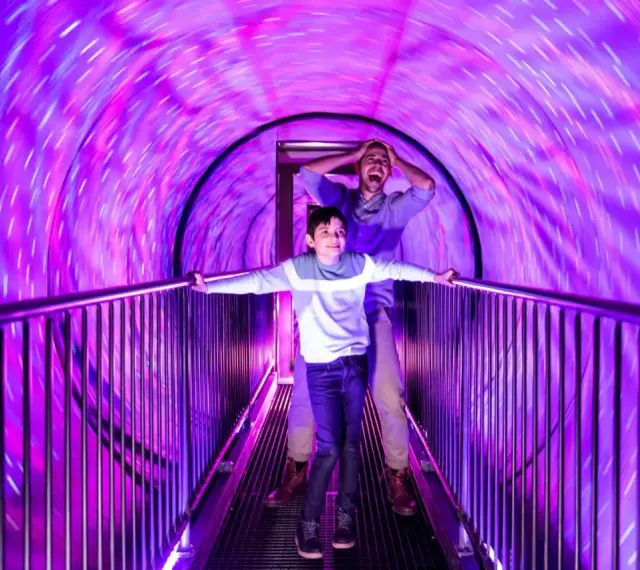 Un garçon et un homme traversent un tunnel lumineux et tourbillonnant dans un musée de l'illusion. Leurs visages expriment la surprise et l'amusement.