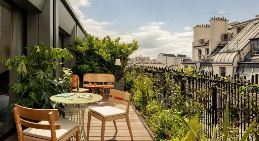 Aménagement balcon 10m2 : table ronde verte, chaises en bois, banquette en rotin, plantes luxuriantes et vue sur les toits de Paris.