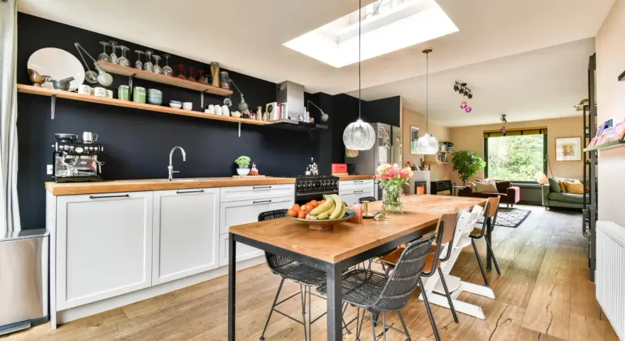Cuisine moderne avec îlot central, parfait pour rénover vieille cuisine. Table en bois, chaises design, et mur noir avec étagères.