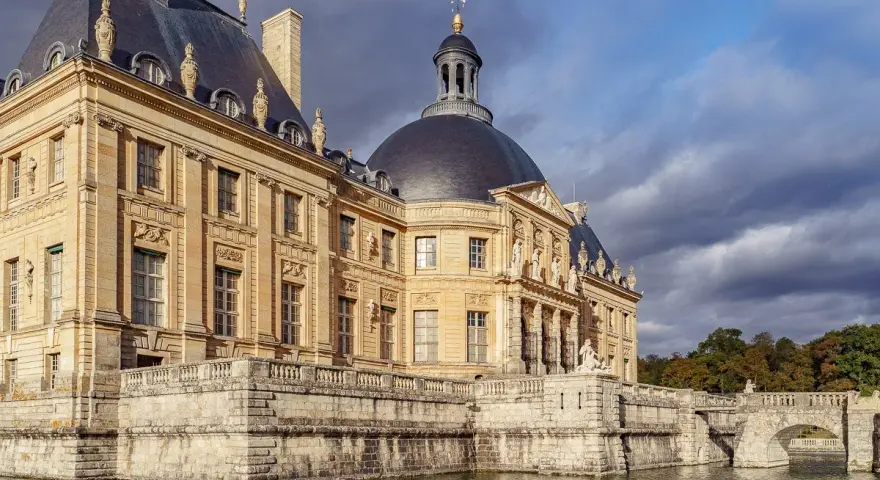 Le château de Vaux-le-Vicomte, majestueux édifice de pierre dorée, se reflète dans les eaux calmes, sous un ciel orageux.