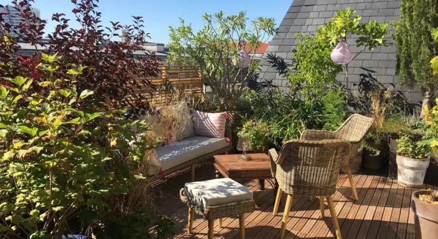 Aménagement terrasse jardin : salon de jardin en rotin, banquette confortable, table basse en bois, plantes luxuriantes et lanternes suspendues sous un ciel bleu.