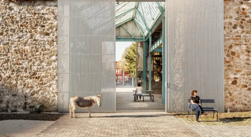 Un âne se tient devant la ferme de la Villette, une structure moderne contrastant avec un mur de pierre ancien. Deux personnes profitent du soleil sur des bancs.