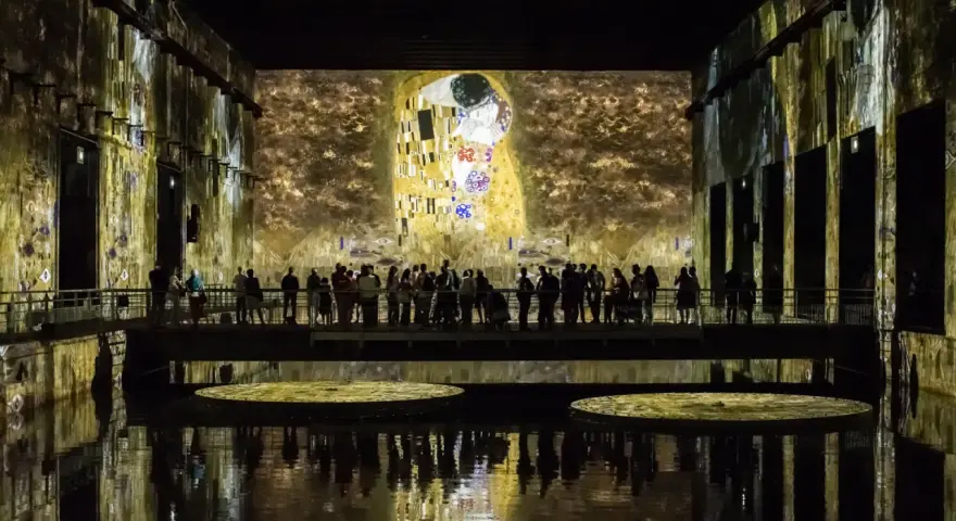 Un bassin des lumières projette "Le Baiser" de Klimt sur les murs d'une salle immersive. Des visiteurs contemplent l'œuvre depuis une passerelle.