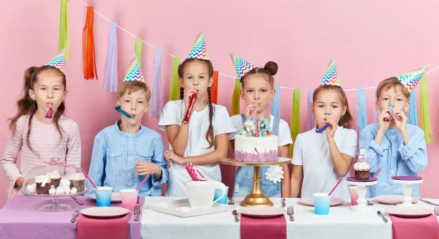 Six enfants joyeux célèbrent un anniversaire 11 ans, portant des chapeaux de fête et soufflant dans des cotillons devant un gâteau décoré.