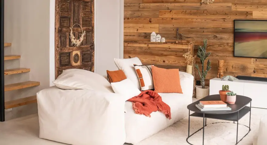 Un canapé blanc moelleux, des coussins orange et un plaid créent une deco petit salon cosy. Une porte ancienne et un mur en bois ajoutent du caractère.