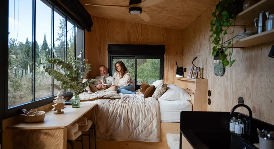 Un couple lit dans un lit douillet, profitant de l'aménagement ingénieux d'une tiny house avec vue sur la nature.