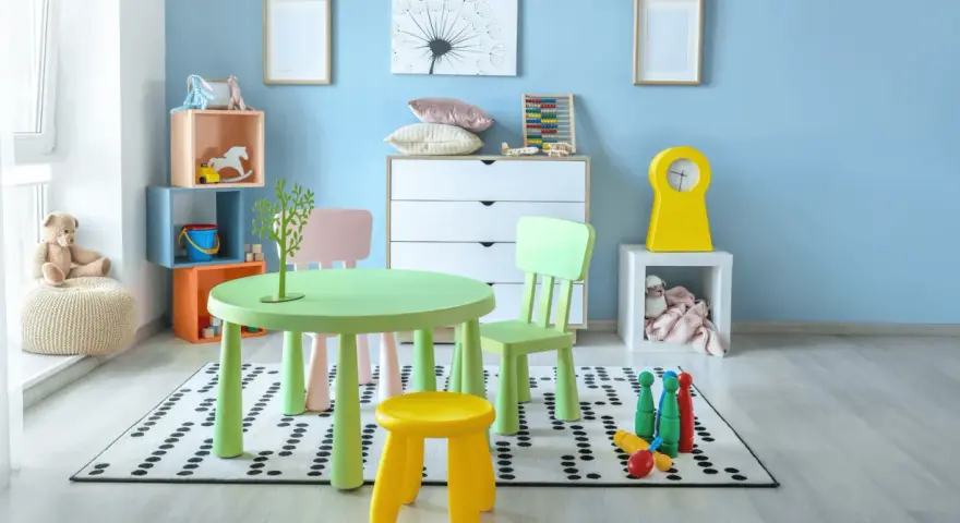 Salle de jeux lumineuse avec table verte, chaises colorées, jouets et peluche sur fond bleu clair.