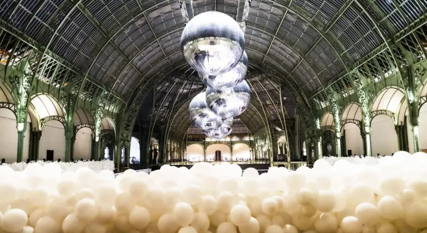 Installation de ballons blancs dans le Grand Palais, Paris. Des sphères argentées flottent sous la verrière, évoquant un musée des ballons.
